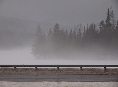 [The guardrail for the bridge in the foreground is clear, but the rest of the image is misty. Plowed dirty snow covers the lower half of the guardrail posts, but the road is just wet. There is a jut of land with evergreens sticking into the lake. The lake itself is covered with white snow and the air just above the water is quite thick with fog. Although the area above the trees is a bit clearer, the visibility is low.]